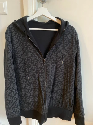Svart MK zip-hoodie - Snygg svart hoodie från Michael Kors med dragkedja och huva. Hoodien har ett diskret MK-mönster över hela tyget, svarta muddar och dragsko i huvan. Perfekt för en stilren och avslappnad look. (Pris går att diskutera) !?
