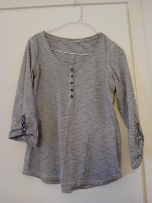 Grå randig blus med knappar - Grå blus med tunna ränder och trekvartsärm. Rund halsringning och knappdetaljer framtill. Ärmarna har knapp och slejf för uppvik. Perfekt till jeans eller kjol.