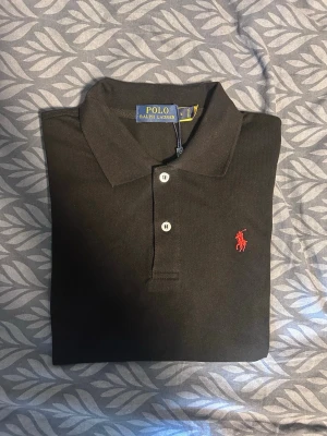Svart pikétröja från Polo Ralph Lauren - Klassisk svart pikétröja från Polo Ralph Lauren med röd broderad logga på bröstet och vita knappar. Tröjan har krage och är tillverkad i bomull, perfekt för en stilren look. Storleken är M men passar S. Pris kan diskuteras.