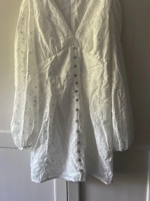 Vit broderad kortklänning med knappar och lång puffärm. Stl M - Superfin vit kortklänning med broderade blommor, v-ringning och långa puffärmar. Klänningen har dekorativa knappar framtill och är tillverkad i ett lätt tyg som ger en romantisk känsla. Helt ny då vi köpte en lika men mindre stl. Denna är i stl M och den är normal i storleken. Mycket lik Ad___e klänning. Passar mycket bra till student skolavslutning eller finare högtider. 