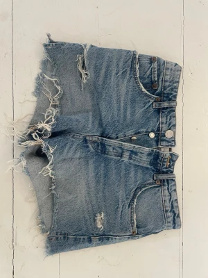 Blå denimshorts med fransar från Zara - Säljer ett par blå denimshorts från Zara med råa, fransiga kanter och slitna detaljer. Klassisk femficksmodell med hög midja och knappgylf. 