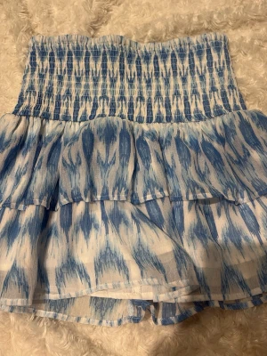 Blåvit volangkjol med smock - Supersöt kort kjol i blått och vitt med smock i midjan och volangdetaljer. Kjolen har ett abstrakt mönster och är gjord i ett lätt, luftigt material som passar perfekt till varma dagar.