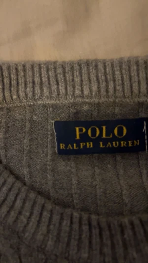 Grå kabelstickad tröja Polo Ralph Lauren/priset kan diskuteras  - Kabelstickad grå tröja från Polo Ralph Lauren med rund halsringning och långa ärmar. Klassisk design med ribbade muddar och diskret broderad logga på bröstet. Perfekt för en stilren och avslappnad look.