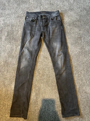 Nudie jeans grim tim - Säljer ett par gråa skinny jeans från Nudie Jeans med klassisk femficksdesign och diskreta slitningar. Jeansen har smal passform och är tillverkade i stretchigt denimtyg för extra komfort. Perfekta för dig som gillar en stilren och modern look.
