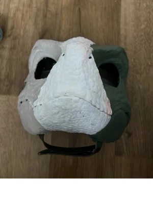 Dinosauriemask i plast - Cool dinosauriemask i grönt och vitt med stor nos och tydliga detaljer. Masken har öppningar för ögonen och ett elastiskt band för att sitta bekvämt på huvudet. Perfekt för maskerad eller cosplay.