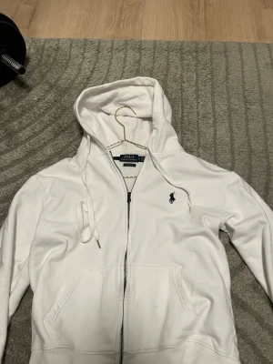 Vit hoodie från Polo Ralph Lauren - Snygg vit hoodie med dragkedja från Polo Ralph Lauren. Klassisk design med huva och dragsnören, två fickor framtill och den ikoniska broderade loggan på bröstet. Perfekt för en clean och stilren look.