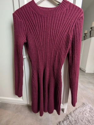 Vinröd ribbad stickad klänning - Mysig vinröd stickad klänning med ribbat mönster och lång ärm. Klänningen har rund halsringning och är figurnära upptill med lätt utställd kjol. Perfekt för kyligare dagar och enkel att styla med boots eller sneakers.