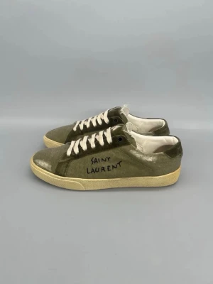 Saint Laurent gröna sneakers - Snygga gröna sneakers från Saint Laurent med handskriven logga på sidan. Skorna har vita snören, beige sula och ovandel i canvas med mockadetaljer. Låg modell som passar perfekt till streetwear och avslappnade outfits.