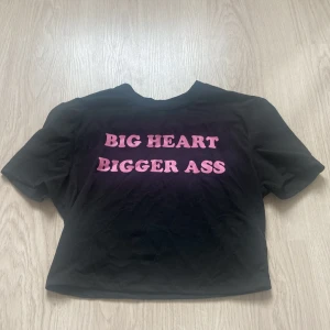 Svart croppad t-shirt med text - Svart croppad t-shirt med rosa texten 'BIG HEART BIGGER ASS' framtill. T-shirten har korta ärmar och en tight passform, perfekt för att göra ett statement. Materialet känns mjukt och bekvämt mot huden.