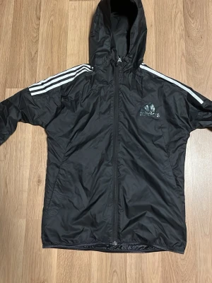 Svart vindjacka från Adidas - Svart vindjacka från Adidas med huva och dragkedja framtill. Klassiska vita ränder längs axlarna och Adidas-logga på bröstet. Tillverkad i lätt, vindtätt material som passar perfekt för blåsiga dagar. Sportig och stilren design.