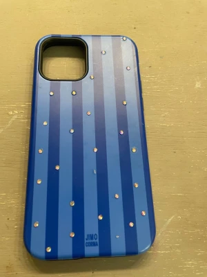 Blått randigt mobilskal med stenar Ipone 12 - Snyggt mobilskal i blå plast från Jimo Corma med vertikala ränder i olika blå nyanser och små glittrande stenar på baksidan. Skalets insida är svart och ger bra skydd till mobilen. Perfekt för dig som vill ha ett unikt och stilrent skal. Ipone12!
