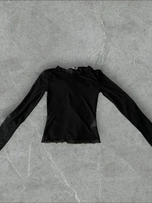 Svart långärmad topp i mesh - Snygg svart långärmad topp i meshmaterial med figurnära passform. Toppen har rund halsringning och vågiga kanter vid ärmslut och nederkant. Perfekt att styla lager på lager eller som statement-piece.