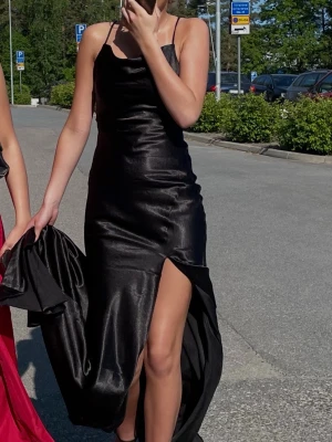 Svart långklänning med slits - Snygg svart långklänning i glansigt material med tunna axelband och hög slits på ena sidan. Klänningen har en elegant, figurnära passform och en enkel men stilren design som passar perfekt till speciella tillfällen. Endast använd 1 gång, nypris 1300