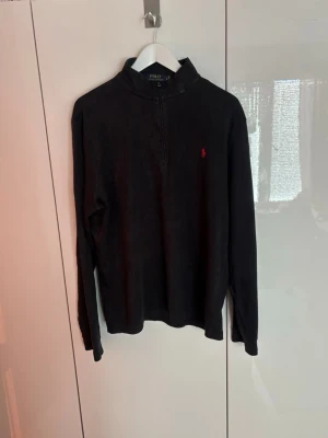 Svart halvzip från Polo Ralph Lauren - Svart långärmad tröja från Polo Ralph Lauren med halv dragkedja och ståkrage. Klassisk röd logga broderad på bröstet. Tillverkad i mjukt material som känns skönt mot huden. Perfekt för en clean och stilren look.