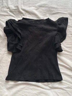 Svart topp med volangärmar - Snygg svart ribbad topp från Gina Tricot med volangdetaljer på ärmarna. Toppen har rund halsringning och är tillverkad i mjukt material som sitter bekvämt. Perfekt för dig som vill ha en trendig och stilren look.