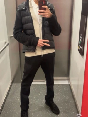 Svart pufferjacka med huva - Snygg svart pufferjacka med quiltad front och stickade ärmar. Jackan har huva med dragsko och dragkedja framtill. Perfekt för kyliga dagar och ger en modern look. Passar dig som vill ha en stilren och bekväm jacka.