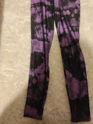 Lila och svarta tie dye leggings - Säljer ett par stretchiga leggings med coolt lila och svart tie dye-mönster. De är i ett mjukt, följsamt material som sitter snyggt på benen. Perfekta för dig som vill sticka ut lite extra med din outfit!