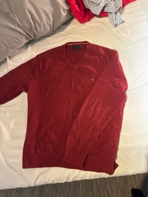 Röd V-ringad stickad tröja Tommy Hilfiger - Snygg röd stickad tröja från Tommy Hilfiger med V-ringning och diskret logga på bröstet. Tröjan har långa ärmar och ribbade muddar vid ärmslut och nederkant. Perfekt för lager på lager och enkel att matcha. Den är tillverkad i Kashmir så den är riktigt skön!