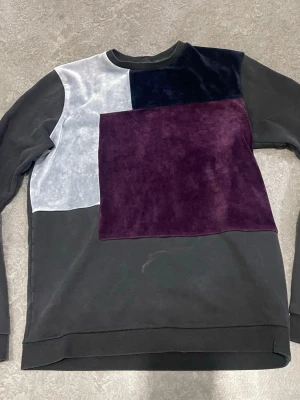 Patchwork sweatshirt från Vichy - Unik sweatshirt från Vichy med patchwork-design i svart, lila och ljusgrå sammet. Tröjan har rund halsringning och en cool badge på ärmen. Materialmix med både bomull och sammetsdetaljer för extra stilpoäng. Perfekt för dig som vill sticka ut.