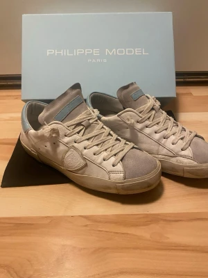 Philippe Model vita sneakers - Snygga vita sneakers från Philippe Model med ljusblå detaljer på plösen och hälen. Skorna har en klassisk låg siluett, vita skosnören och en ikonisk sköldlogga på sidan. Tillverkade i läder och mocka för en exklusiv känsla.