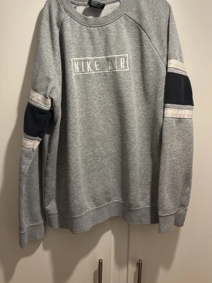 Grå Nike Air sweatshirt - Snygg grå sweatshirt från Nike Air med vitt tryck på bröstet. Tröjan har svarta och vita detaljer på ärmarna och en avslappnad passform. Perfekt för en sportig och casual stil.