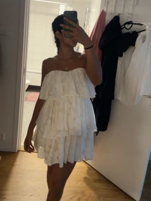 Vit volangklänning axelbandslös från fashion nova - Säljer en söt vit axelbandslös klänning med flera lager volanger och blommigt mönster. Klänningen är kort och har en luftig passform.