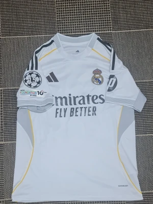 Real Madrid matchtröja Adidas vit - Säljer en vit Real Madrid matchtröja från Adidas med klubbmärke, Champions League-märke och UEFA Foundation-märke på ärmen. Tröjan har grå och gula detaljer, samt 'Emirates Fly Better' tryckt på bröstet. Tillverkad i lätt och ventilerande material.