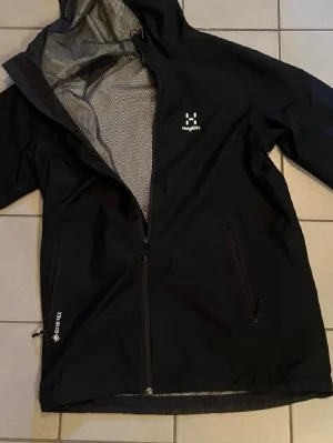 Svart regnjacka från Haglöfs Gore-Tex - Svart regnjacka från Haglöfs med Gore-Tex-material som håller dig torr. Jackan har huva, hel dragkedja framtill, två sidofickor med dragkedja och meshfoder på insidan för extra ventilation. Perfekt för regniga dagar och outdoor-äventyr.