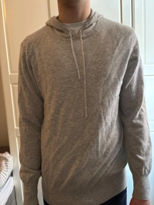 Kashmirhoodie från Soft Goat - Mjuk och enkel ljusgrå hoodie med huva och dragsnören. Tillverkad i ett skönt stickat material som känns bekvämt mot huden. Perfekt för en avslappnad stil och passar till det mesta. Långa ärmar och ribbade muddar vid ärmslut och nederkant.