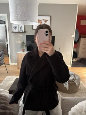 Svart omlottkappa med knytskärp - Stilren svart kappa i omlottmodell med brett slag och knytskärp i midjan. Kappan har lång ärm och rak passform som ger en snygg siluett. 