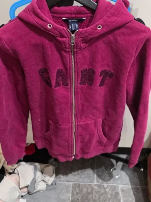 Vinröd hoodie med dragkedja från GANT - Säljer en vinröd hoodie från GANT med dragkedja framtill och broderad logga på bröstet. Hoodien har två fickor fram och en skön huva. Tillverkad i mjuk bomullsblandning som känns riktigt bekväm. Perfekt för dig som vill ha en stilren och avslappnad look.