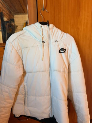 Vit pufferjacka från Nike med huva - Säljer en vit pufferjacka från Nike med huva och dragkedja framtill. Jackan har svarta detaljer som Nike-logga på bröstet och snören i huvan. Perfekt för kalla vinterdagar och riktigt skön att bära. Fodrad för extra värme och har en sportig look. Priset kan diskuteras vid snabb affär.