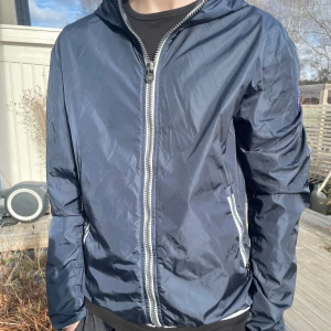 Mörkblå vindjacka från Colmar - Snygg mörkblå vindjacka från Colmar med vit dragkedja och två fickor framtill. Jackan har huva och Colmar-logga på ärmen. Tillverkad i lätt, glansigt material som står emot vind. Nypris 1499kr. Skicket utanpå är så gott som ny, MEN skicket innanför jackan är väldigt slitet därav de de billigare priset