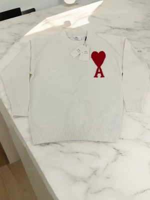 Vit stickad tröja Ami Paris hjärta - Vit stickad tröja från Ami Paris med röd hjärt- och A-logga på bröstet. Tröjan har rund hals, ribbade muddar och nederkant. Klassisk och stilren design som passar till mycket. Materialet är mjukt och bekvämt.