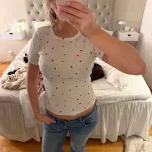 Vit t-shirt med röda hjärtan - Supersöt vit t-shirt med små röda hjärtan över hela. T-shirten har rund halsringning och korta ärmar. Perfekt för dig som gillar gulliga detaljer och en enkel men charmig stil.