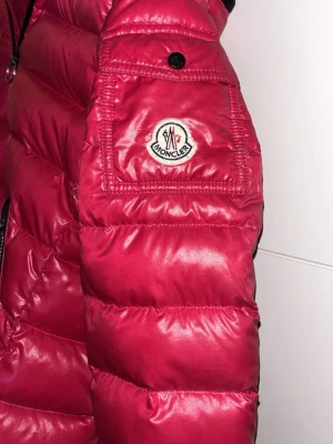 Röd dunjacka från Moncler - Röd/ljusröd dunjacka från Moncler med glansigt yttertyg, svart dragkedja och svarta detaljer vid fickorna. Jackan har en huva och en patch med Moncler-loggan på ärmen. Puffig modell som håller dig varm under vintern och ger en riktigt trendig vibe. Barnstorelk 146/152