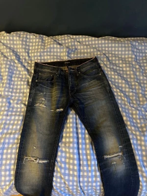 Mörkblå slitna jeans med hål - Säljer ett par mörkblå jeans med slitningar och hål på båda benen. Jeansen har en klassisk femficksmodell och är tillverkade i denim med stretch. Perfekta för dig som gillar en avslappnad och trendig stil.
