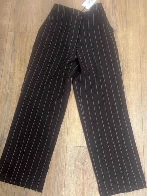 Svarta kostymbyxor med vita ränder - Snygga svarta kostymbyxor med vita vertikala ränder och rak passform. Byxorna har hög midja och klassisk look, perfekta för en stilren outfit. Materialet känns mjukt och följsamt, vilket ger en bekväm känsla hela dagen.