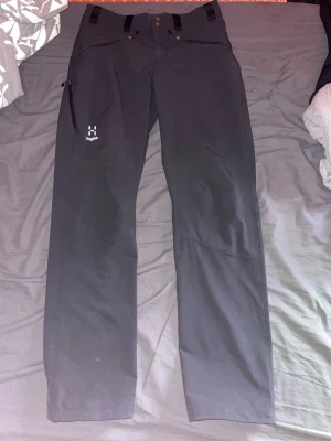 Svarta softshellbyxor från Haglöfs - Säljer ett par svarta softshellbyxor från Haglöfs med raka ben, dragkedja fram och praktisk ficka med dragkedja på sidan. Byxorna har bälteshällor och diskret Haglöfs-logga på låret. Perfekta för chill dagar eller när du vill ha det bekvämt.