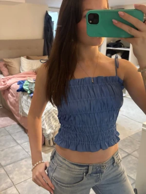 Blå smockad linnetopp med tunna band - Säljer en blå croppad linnetopp med smockad design och tunna axelband som knyts upptill. Toppen har en rynkad effekt och är perfekt för varma dagar. Enkel och trendig stil som passar till jeans eller kjol.