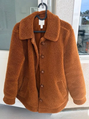Brun teddyjacka med stora knappar - Mysig brun teddyjacka med stora knappar framtill och två fickor. Jackan har en klassisk krage och är gjord i fluffigt teddy-material som håller dig varm. Perfekt för kyliga dagar och ger en trendig look. Passar dig som gillar oversized och avslappnad stil. Passar mer till strl 38/40