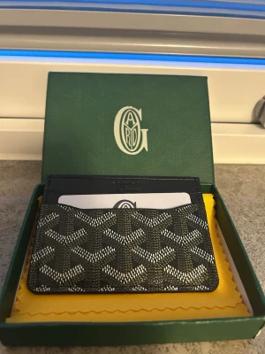 Svart kortfodral - Snyggt svart kortfodral med det klassiska vita och gråa mönstret. Tillverkat i canvas och läder, perfekt för kort och ID. Kommer i original grön box och med gul putsduk. Stilren och lyxig accessoar för dig som gillar exklusiva detaljer.