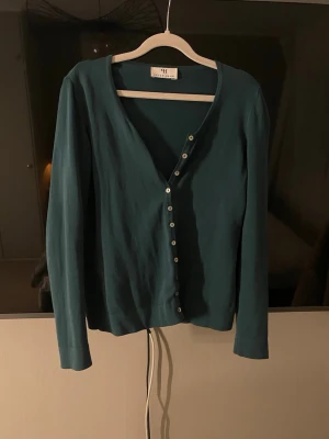 Mörkgrön kofta från Peter Hahn - Stilren mörkgrön kofta från Peter Hahn med v-ringning och knäppning framtill. Långa ärmar och smal passform, perfekt att slänga över en topp. Klassisk design med små vita knappar och mjukt material.