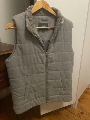 Grå väst från Dressmann - Snygg grå quiltad väst från Dressmann med dragkedja framtill och två fickor. Västen har en hög krage i manchestertyg och är vadderad för extra värme. Perfekt att slänga över en hoodie eller tröja för en avslappnad look. Pris går att disskutera vid snabb affär :)