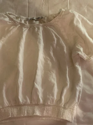 Vit blus med puffärm och resår - Söt vit blus med korta puffärmar och bred resår i nederkant. Blusen har rund halsringning och är gjord i ett lätt tyg som ger en luftig känsla. Perfekt att styla med jeans eller kjol för en fräsch look.