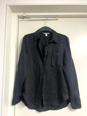 Svart linneskjorta från H&M - Stilren svart skjorta från H&M i 100% linne. Skjortan har klassisk krage, lång ärm och knappar framtill samt en bröstficka. Perfekt för en avslappnad men ändå snygg look. Passformen är loose och materialet känns svalt och luftigt.
