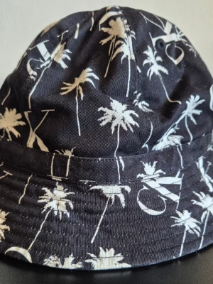 Svart bucket hat  - Säljer en svart bucket hat . Ena sidan har ett vitt palm- och strandmönster, medan andra sidan är helsvart med en stor CK-logga framtill.  Passar mer för barn