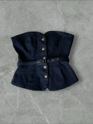 Mörkblå denimkorsett med bälte - Trendig mörkblå korsett i denim med markerad midja och avtagbart bälte. Framsidan har dekorativa metallknappar och korsetten har en lätt utsvängd form nedtill. Perfekt för att skapa en edgy look med jeanskänsla.