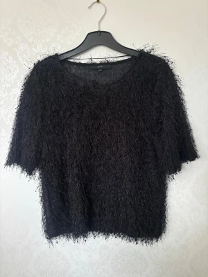 Svart fluffig stickad tröja - Säljer en svart stickad tröja med fluffig, lurvig textur och rund hals. Tröjan har kortare ärmar och en lös passform som ger en avslappnad look. Perfekt för dig som gillar trendiga och unika plagg i garderoben.