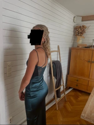 Mörkgrön balklänning - Snygg mörkgrön balklänning i satinliknande material med v-ringad urringning och öppen rygg med spetsdetalj. Klänningen har en slits på sidan och en figurnära passform. Köpt på bubbleroom.